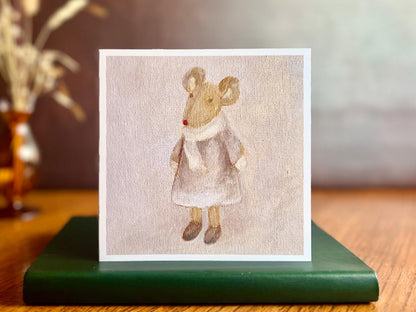 Artisan greeting card featuring a print of an oil painting titled 'Mouse in a Lilac Dress', showing a small brown mouse wearing a lilac dress with a white scarf and mittens, displayed standing on a green leatherette book in a styled lifestyle setting.