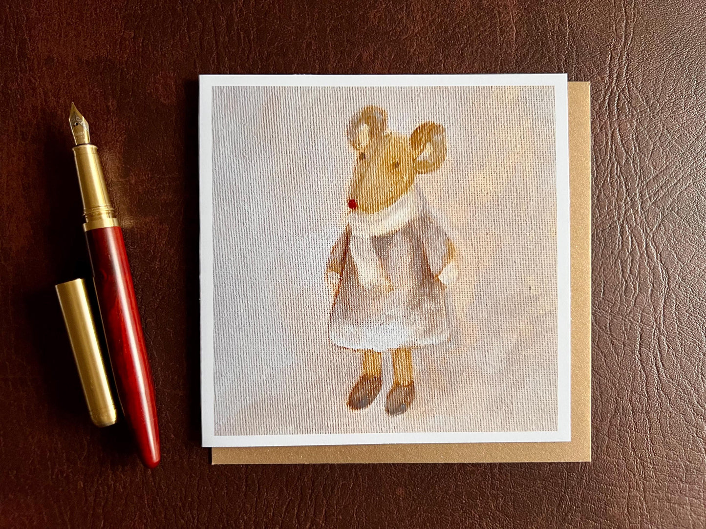 Flat lay image of an artisan greeting card featuring a print of an oil painting titled 'Mouse in Lilac Dress', shown laid flat on a tabletop with its kraft envelope and a fountain pen in a styled composition.