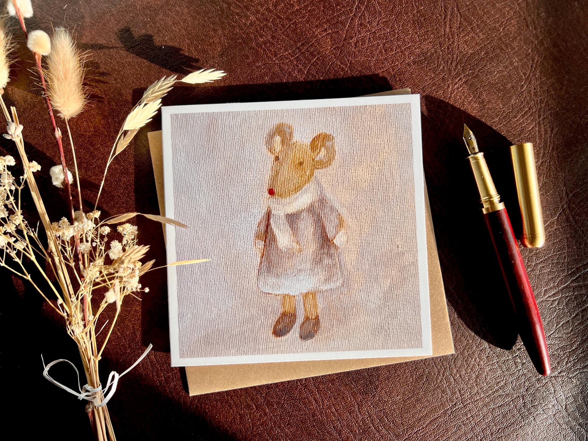 Flat lay image of an artisan greeting card featuring a print of an oil painting titled 'Mouse in Lilac Dress', shown laid flat on a tabletop next to a fountain pen and dried flowers in a styled composition.