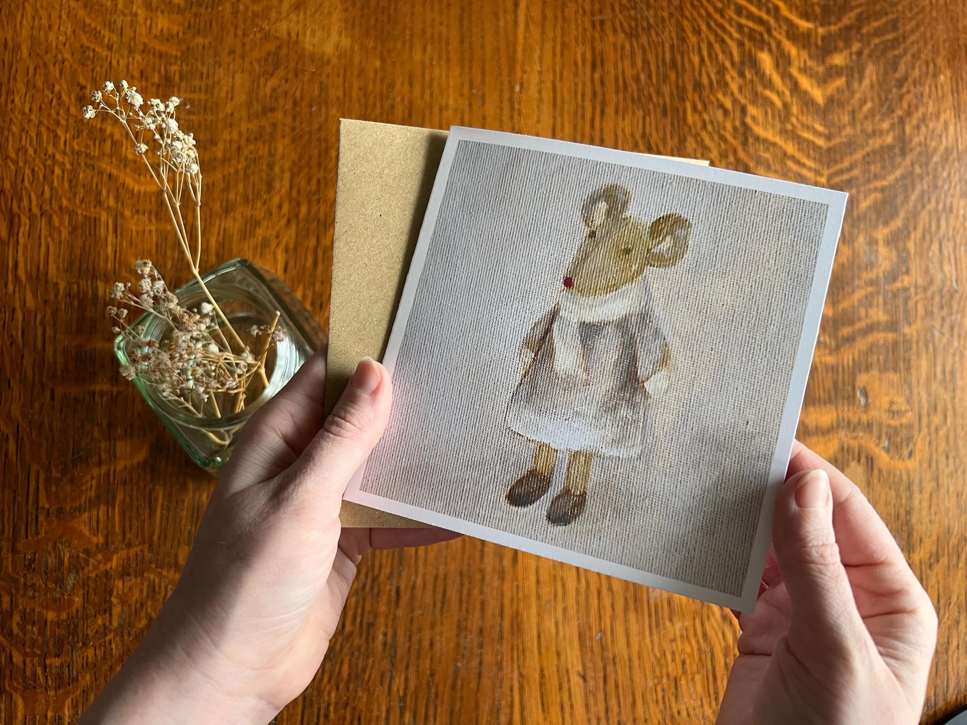 Hands holding an artisan greeting card and its kraft envelope up towards the camera, featuring a print of an oil painting titled 'Mouse in Lilac Dress' showing a small brown mouse wearing a lilac dress with scarf and mittens.