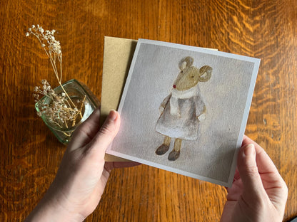 Hands holding an artisan greeting card and its kraft envelope up towards the camera, featuring a print of an oil painting titled 'Mouse in Lilac Dress' showing a small brown mouse wearing a lilac dress with scarf and mittens.