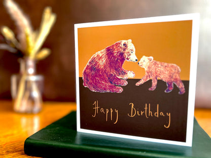 Artisan birthday card featuring a brown bear and cub with 'Happy Birthday' text underneath, displayed upright on a green leatherette book placed on a wooden tabletop in a styled lifestyle setting.