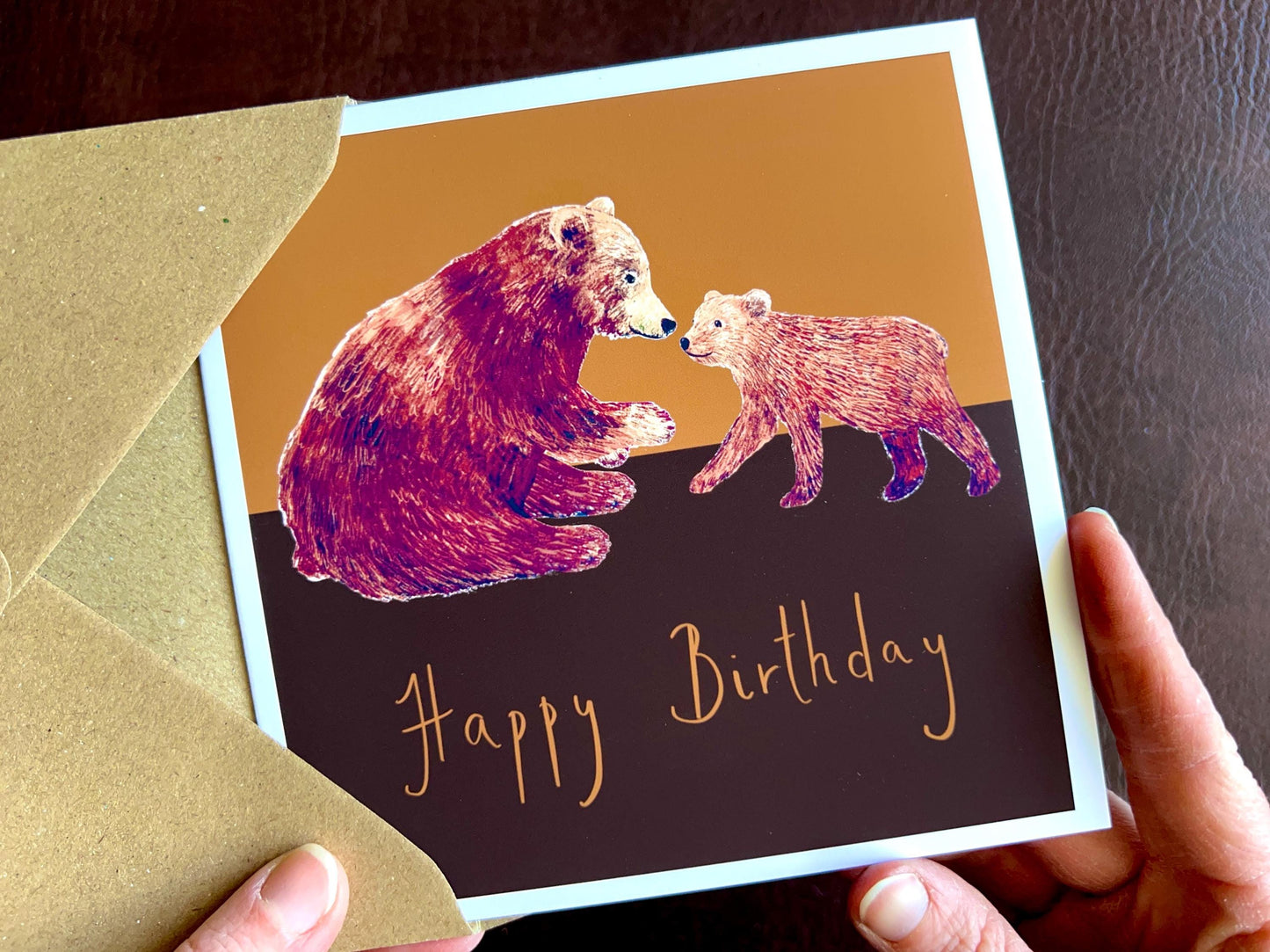 Hands holding an artisan birthday card being removed from a kraft envelope, featuring a brown bear and cub illustration on a warm brown and burnt orange background with 'Happy Birthday' text underneath.