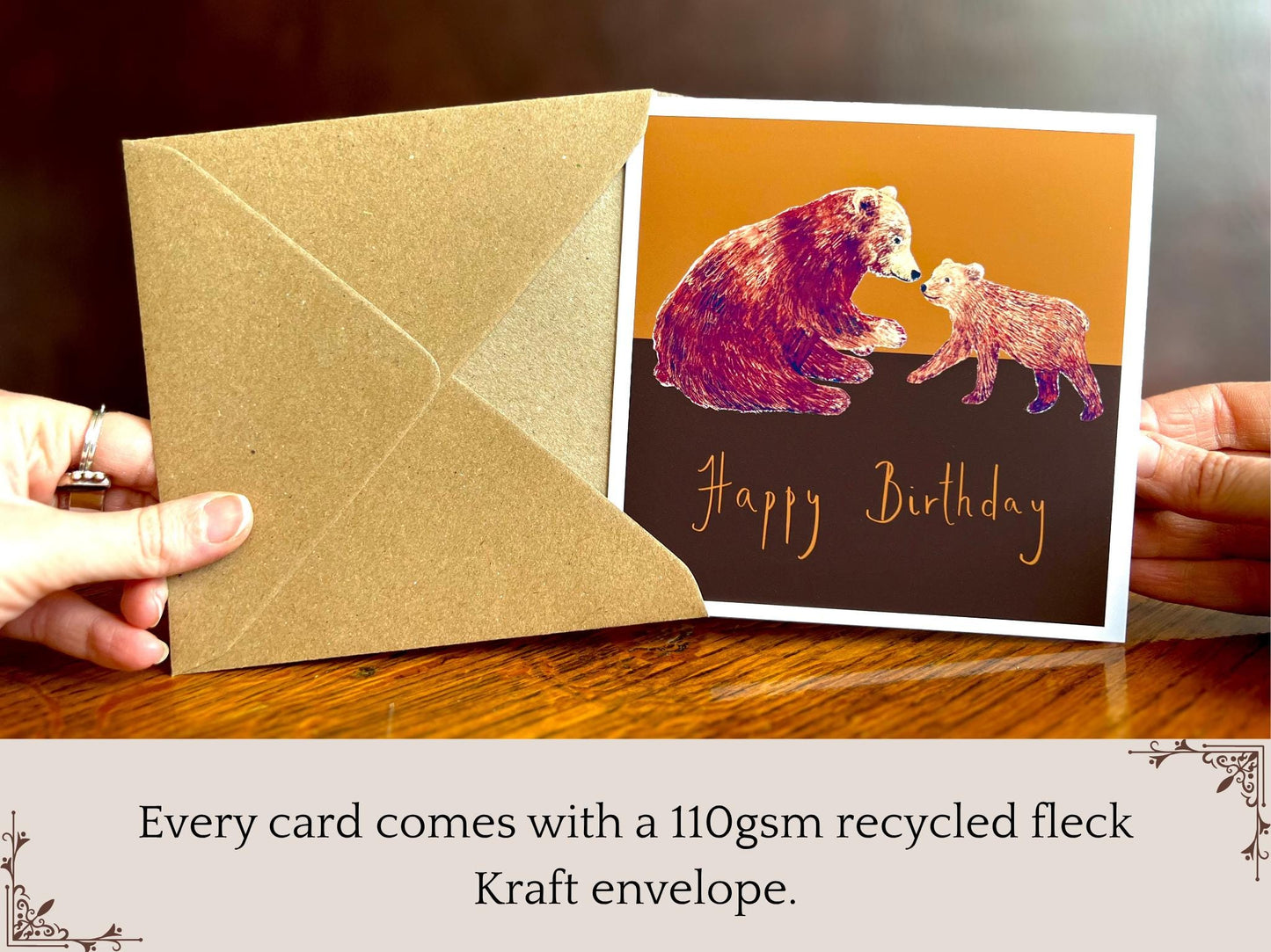 Hands holding an artisan birthday card being removed from a kraft envelope, featuring a brown bear and cub illustration on a warm brown and burnt orange background with 'Happy Birthday' text underneath. Text below the image explains that every card comes with a recycled kraft envelope.