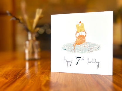 Artisan birthday card featuring a crab holding a sandcastle in clear blue water, shown on a white background. The card displays 'Happy 7th Birthday' beneath the illustration, showing an age personalised version without a custom name.
