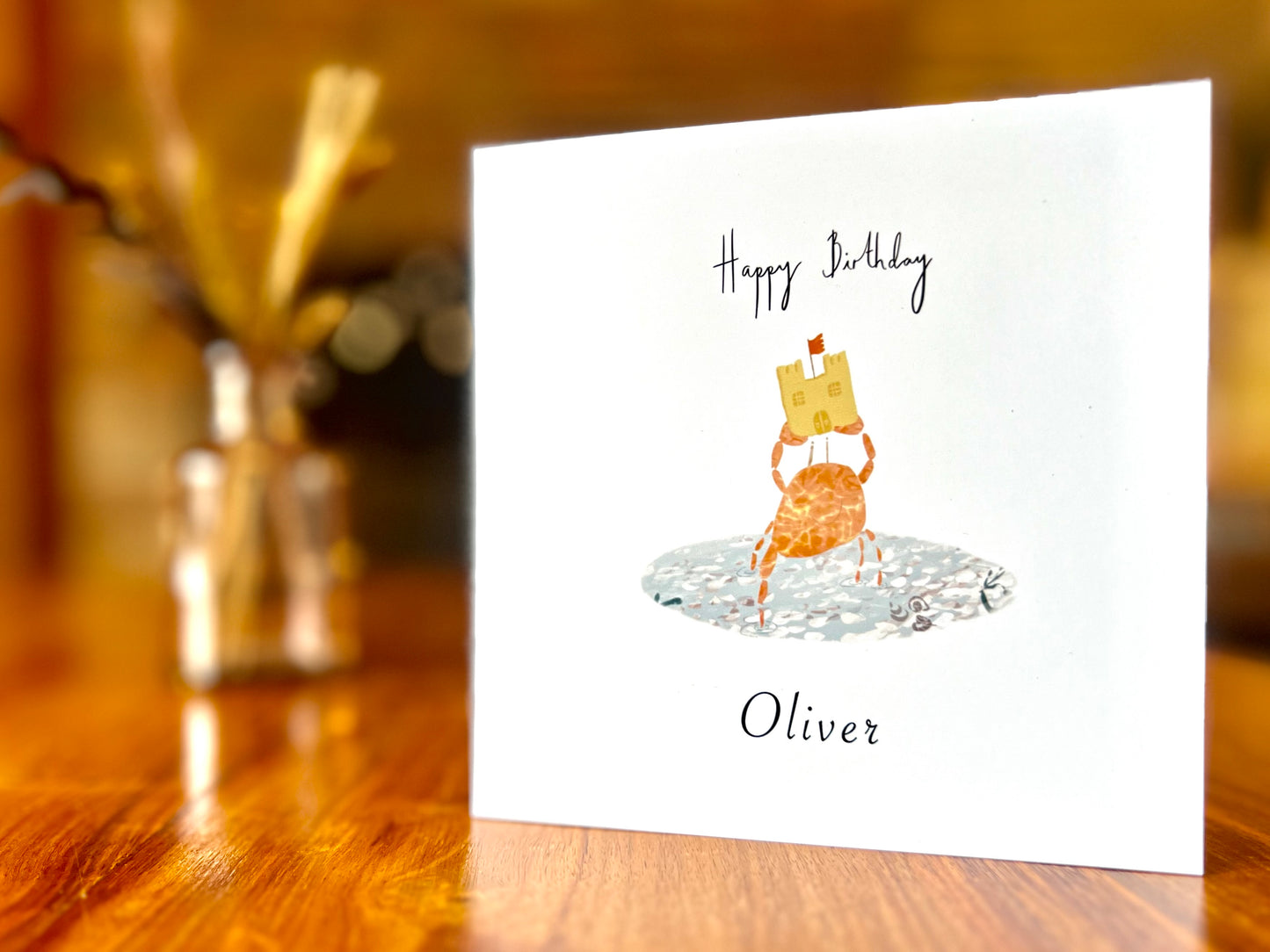 Artisan birthday card featuring a crab holding a sandcastle in clear blue water, displayed upright on a tabletop. The design shows 'Happy Birthday' above the illustration with a custom name underneath, demonstrating personalisation without age.