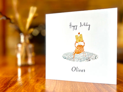 Artisan birthday card featuring a crab holding a sandcastle in clear blue water, displayed upright on a tabletop. The design shows 'Happy Birthday' above the illustration with a custom name underneath, demonstrating personalisation without age.