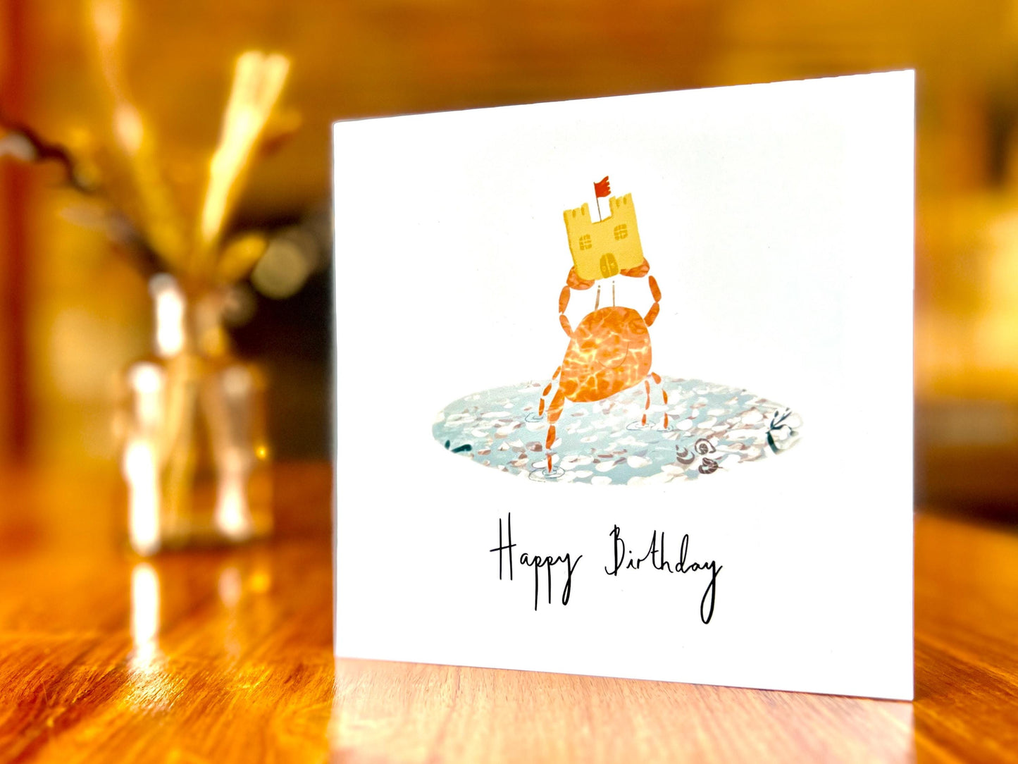 Artisan birthday card featuring a crab holding a sandcastle in clear blue water, shown standing upright on a tabletop. This is the non-personalised version with 'Happy Birthday' written underneath the central illustration on a white background.