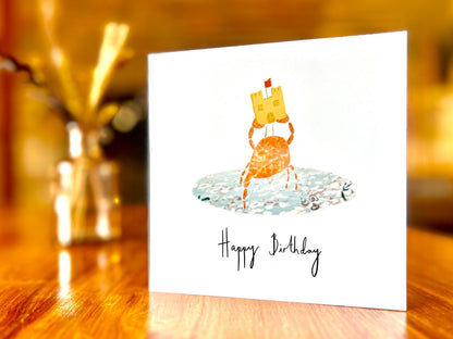 Artisan birthday card featuring a crab holding a sandcastle in clear blue water, shown standing upright on a tabletop. This is the non-personalised version with 'Happy Birthday' written underneath the central illustration on a white background.