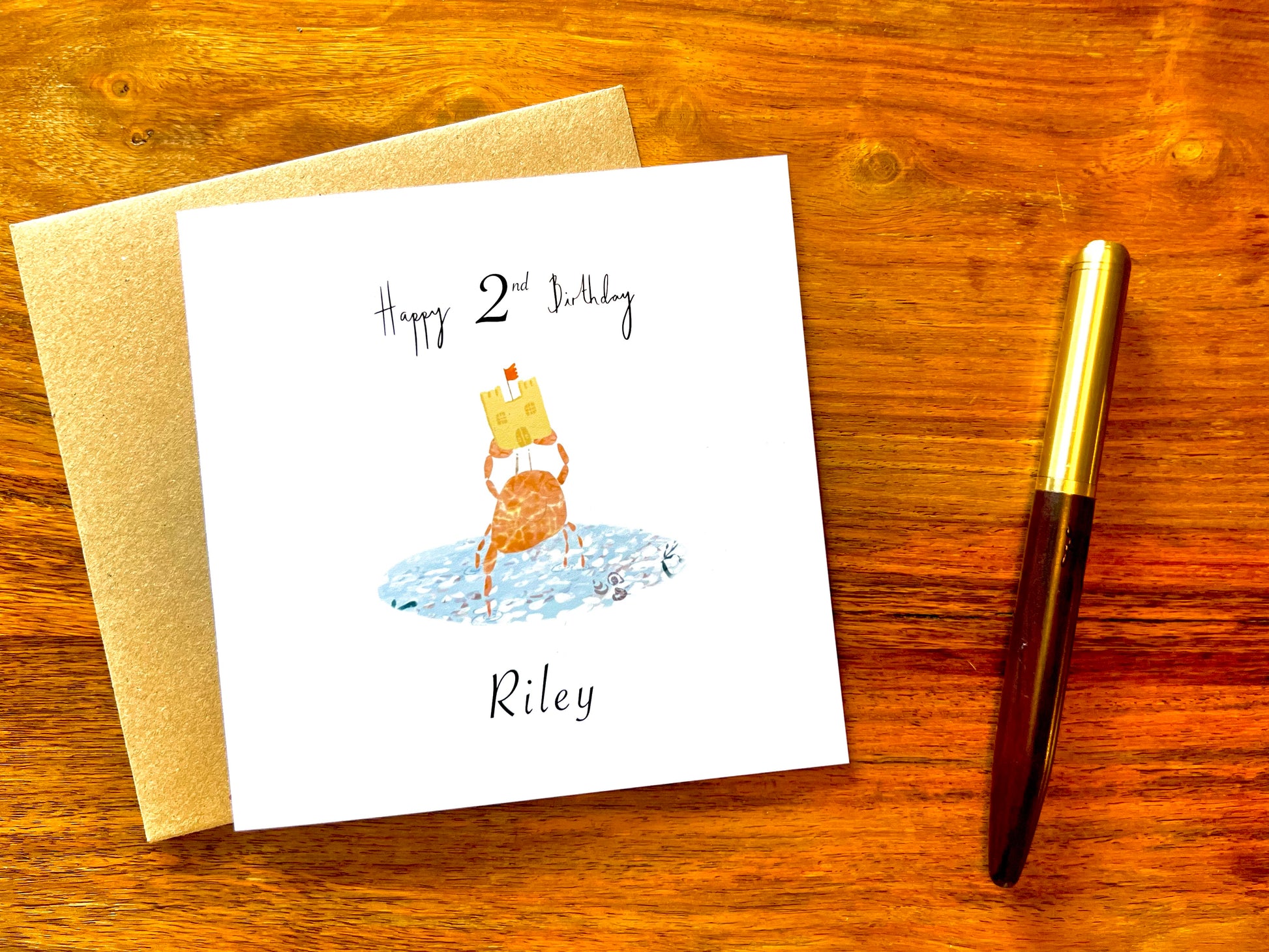 Artisan birthday card featuring a crab holding a sandcastle in clear blue water, shown laid flat on top of a kraft envelope on a tabletop in a styled flat lay composition.
