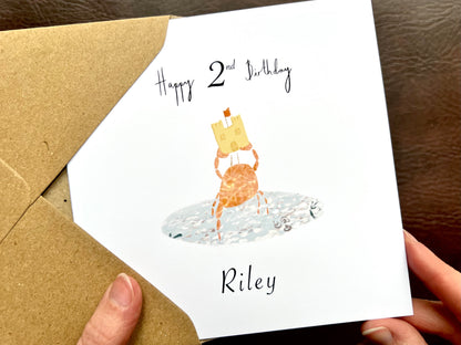 Hands holding an artisan birthday card being removed from a kraft envelope, featuring a fun illustration of a crab holding a birthday sandcastle in clear blue water. The card shows 'Happy 2nd Birthday' above the illustration with a customisable name underneath on a white background.