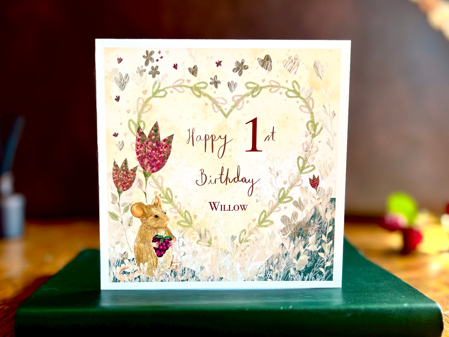 Artisan birthday card featuring an illustrated mouse holding a blackberry with red collage style flowers and a floral heart on a lemon yellow background, shown standing on a green leatherette book on a wooden tabletop. The card reads 'Happy 1st Birthday' with a custom name for personalisation.