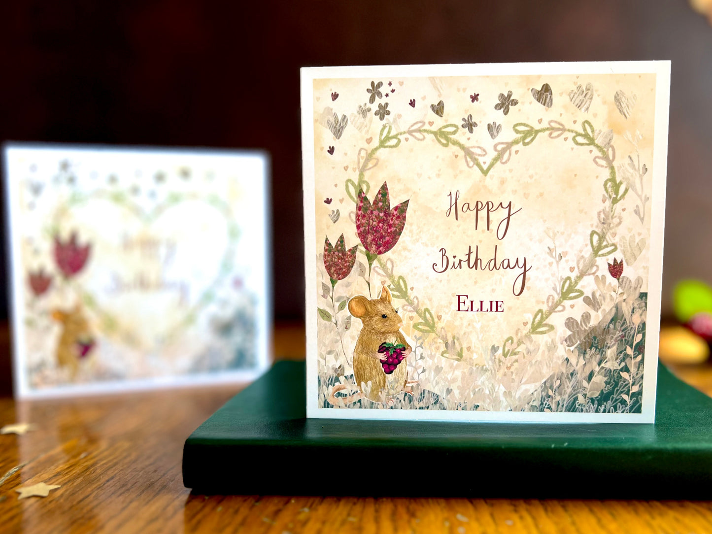 Artisan birthday card featuring an illustrated mouse holding a blackberry with red collage style flowers and a floral heart on a lemon yellow background. The foreground shows the personalised version reading 'Happy Birthday' with a custom name, while the non-personalised version is visible out of focus in the background.