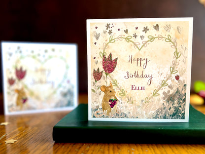 Artisan birthday card featuring an illustrated mouse holding a blackberry with red collage style flowers and a floral heart on a lemon yellow background. The foreground shows the personalised version reading 'Happy Birthday' with a custom name, while the non-personalised version is visible out of focus in the background.