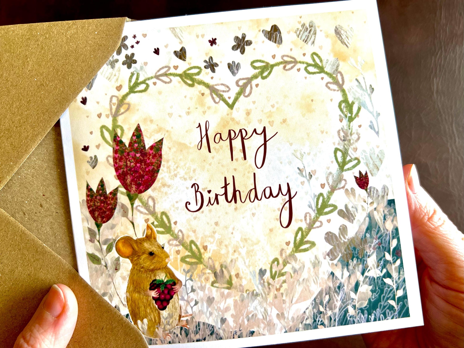 Hands holding an artisan birthday card being removed from a kraft envelope, featuring an illustrated mouse holding a blackberry in the bottom left corner with large red collage style flowers and a floral heart in the centre on a lemon yellow background. The non-personalised version reads 'Happy Birthday' in red handwritten style text.