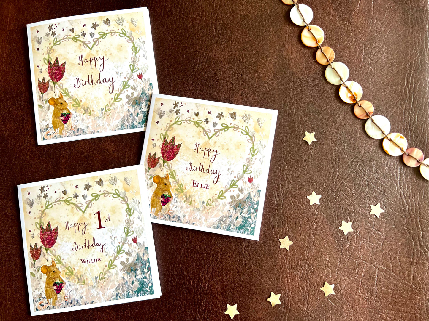 Flat lay image showing three artisan birthday card design variations featuring an illustrated mouse holding a blackberry with red collage style flowers and a floral heart on a lemon yellow background. The versions display 'Happy Birthday', 'Happy Birthday' with a custom name, and 'Happy [age] Birthday' with a custom name for personalisation options.