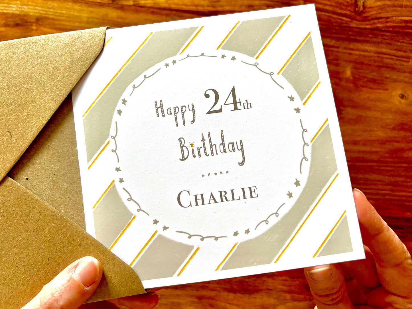 Hands holding an artisan birthday card being removed from a kraft envelope, featuring a pale sage green and white diagonal striped background with a white circle in the centre. The card reads 'Happy 24th Birthday' with a custom name underneath.