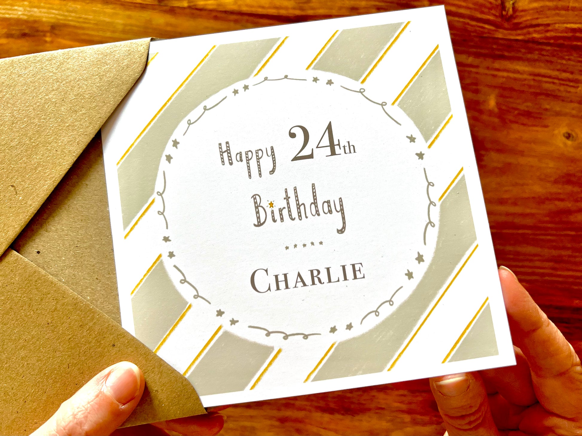 Hands holding an artisan birthday card being removed from a kraft envelope, featuring a pale sage green and white diagonal striped background with a white circle in the centre. The card reads 'Happy 24th Birthday' with a custom name underneath.