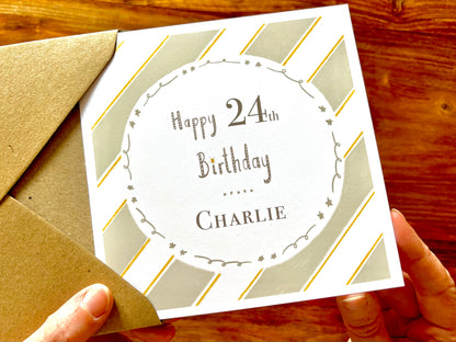 Hands holding an artisan birthday card being removed from a kraft envelope, featuring a pale sage green and white diagonal striped background with a white circle in the centre. The card reads 'Happy 24th Birthday' with a custom name underneath.