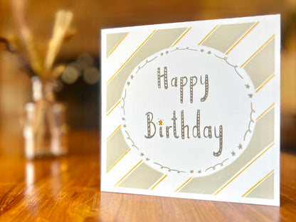 Artisan birthday card with a pale sage green and white diagonal striped background and a white circle in the centre, shown displayed on a wooden tabletop. The card reads 'Happy Birthday' with no customisation added.