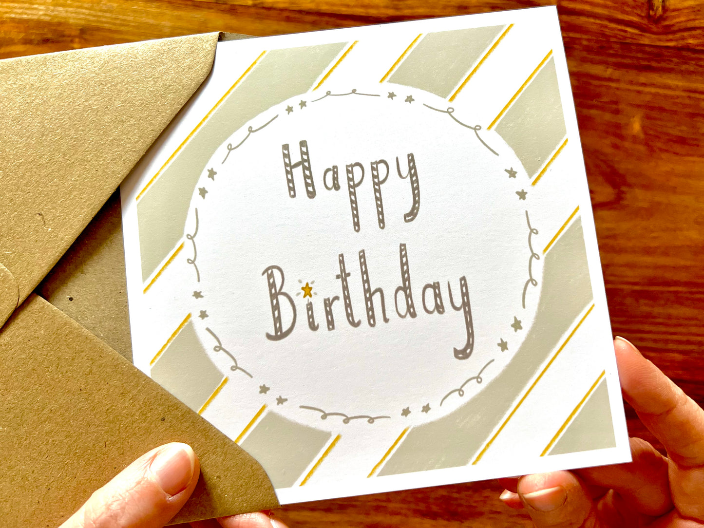 Hands holding an artisan birthday card being removed from a kraft envelope, featuring a pale sage green and white diagonal striped background with a white circle in the centre. The card reads 'Happy Birthday' with no custom name or age added.