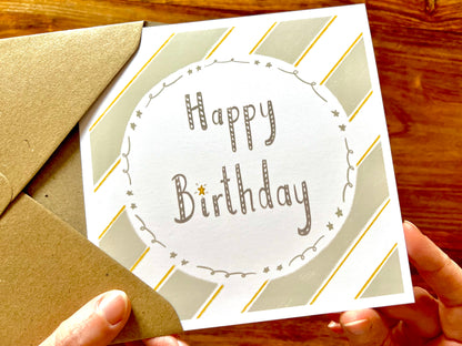 Hands holding an artisan birthday card being removed from a kraft envelope, featuring a pale sage green and white diagonal striped background with a white circle in the centre. The card reads 'Happy Birthday' with no custom name or age added.