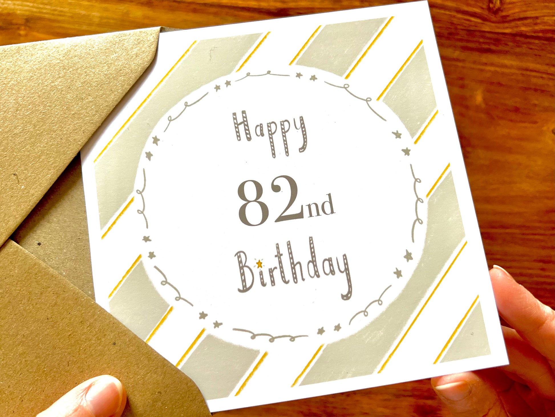 Hands holding an artisan birthday card being removed from a kraft envelope, featuring a pale sage green and white diagonal striped background with a white circle in the centre. The card reads 'Happy 82nd Birthday' and can be customised with age, name, or both.