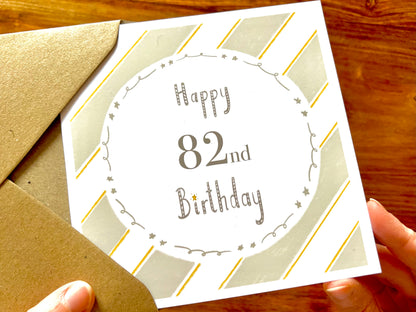 Hands holding an artisan birthday card being removed from a kraft envelope, featuring a pale sage green and white diagonal striped background with a white circle in the centre. The card reads 'Happy 82nd Birthday' and can be customised with age, name, or both.