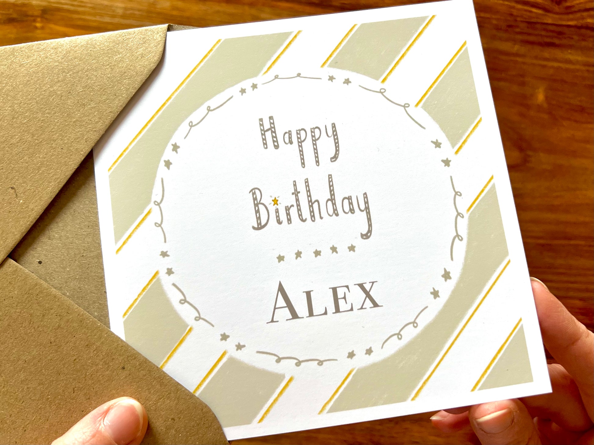 Hands holding an artisan birthday card being removed from a kraft envelope, featuring a pale sage green and white diagonal striped background with a white circle in the centre. The card shows 'Happy Birthday' with a custom name underneath separated by a line of small illustrated stars.