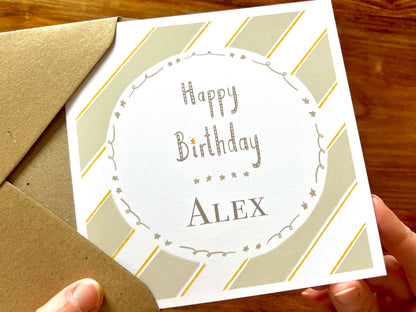 Hands holding an artisan birthday card being removed from a kraft envelope, featuring a pale sage green and white diagonal striped background with a white circle in the centre. The card shows 'Happy Birthday' with a custom name underneath separated by a line of small illustrated stars.