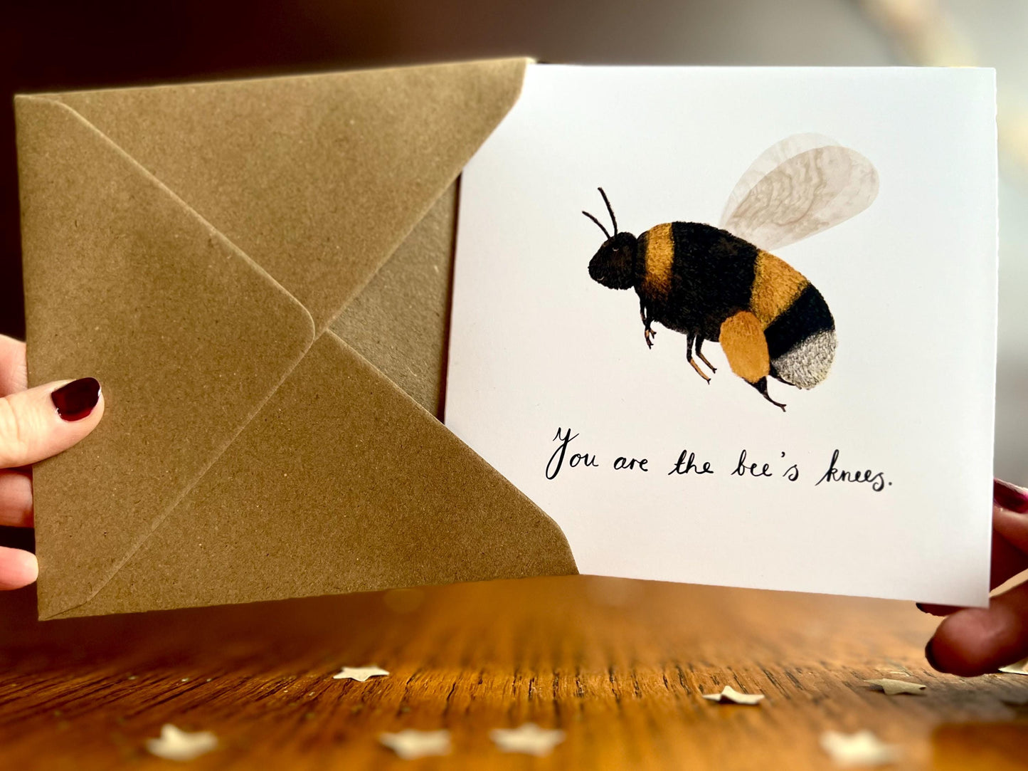 Hands holding an artisan greeting card and its kraft envelope towards the camera, featuring a colourful bumblebee illustration on a white background with the text 'You are the bee’s knees' and a custom name option.