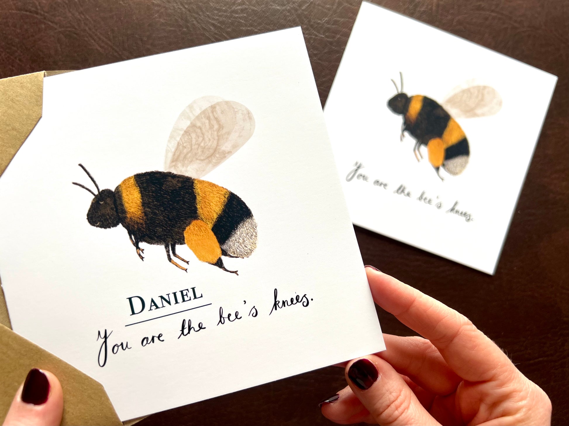 Aerial view of hands holding a personalised artisan greeting card featuring a colourful bumblebee and 'You are the bee’s knees' text with a custom name. In the background, the non-personalised version of the card is laid flat on a tabletop and softly out of focus.