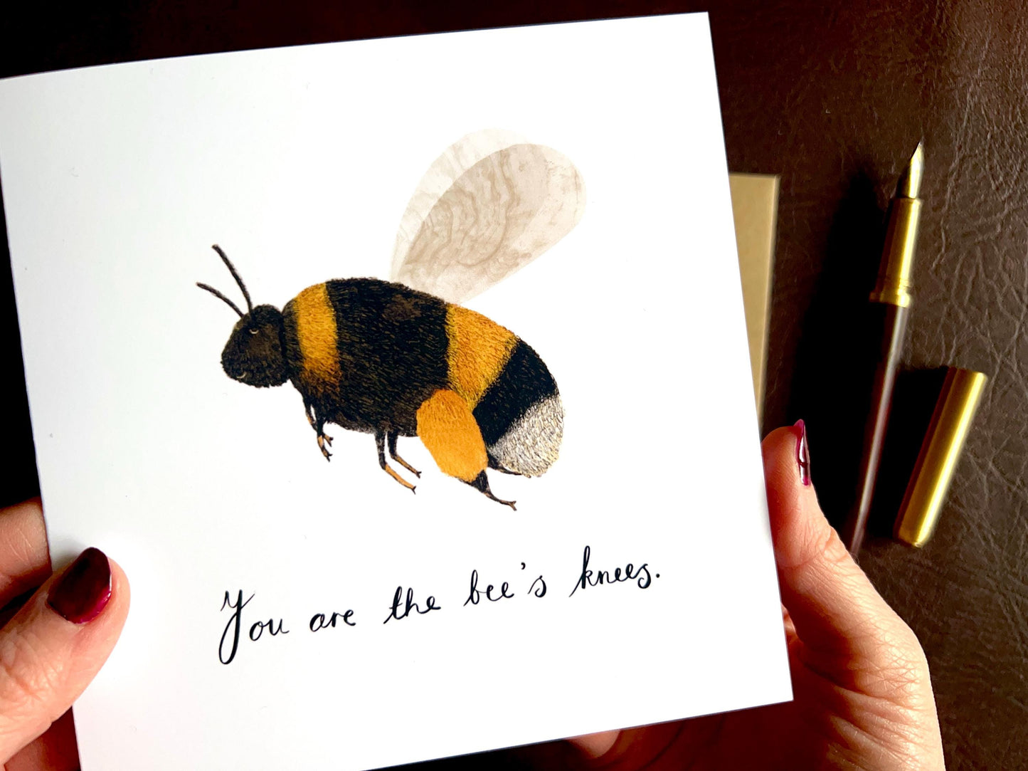 Close up lifestyle image of hands holding an artisan greeting card up towards the camera, featuring a colourful bumblebee illustration on a plain white background with the text 'You are the bee’s knees' and a custom name option.