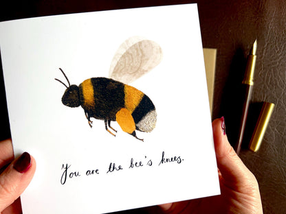 Close up lifestyle image of hands holding an artisan greeting card up towards the camera, featuring a colourful bumblebee illustration on a plain white background with the text 'You are the bee’s knees' and a custom name option.