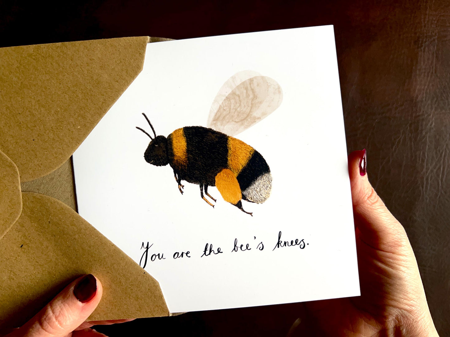 Hands holding an artisan greeting card being removed from a kraft envelope, featuring a colourful bumblebee illustration on a plain white background. The card reads 'You are the bee’s knees' without a custom name, shown as the non-personalised version suitable for general gifting.