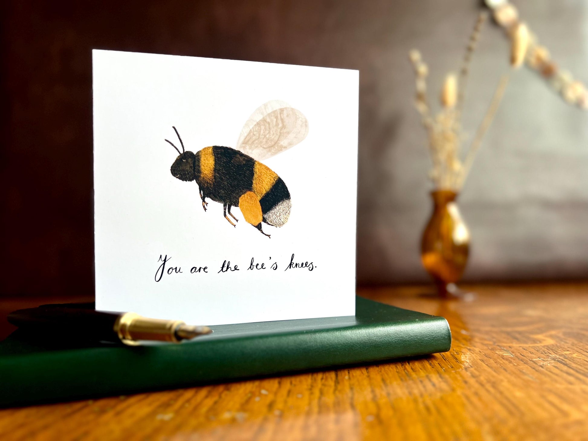Artisan greeting card featuring a colourful bumblebee illustration and the text 'You are the bee’s knees', shown standing upright on a leatherette book placed on a wooden tabletop with a fountain pen in a styled lifestyle setting.