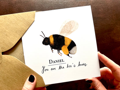 Hands holding an artisan greeting card being removed from a kraft envelope, featuring a colourful bumblebee illustration on a plain white background. The card reads 'You are the bee’s knees' in a handwritten style font with a custom name above for personalisation, suitable for congratulations, good luck or positive wellbeing messages.