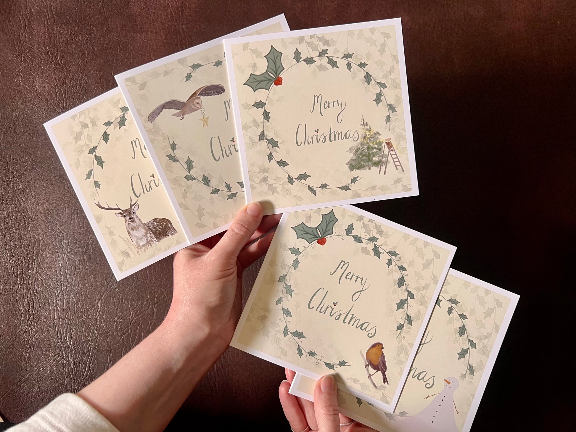 Hands holding a fan-shaped arrangement of five artisan Christmas cards, displaying the full festive collection featuring designs of a mouse decorating a Christmas tree, an owl carrying a star, a snowman, a reindeer, and a robin, all with holly wreath borders and handwritten style 'Merry Christmas' text.