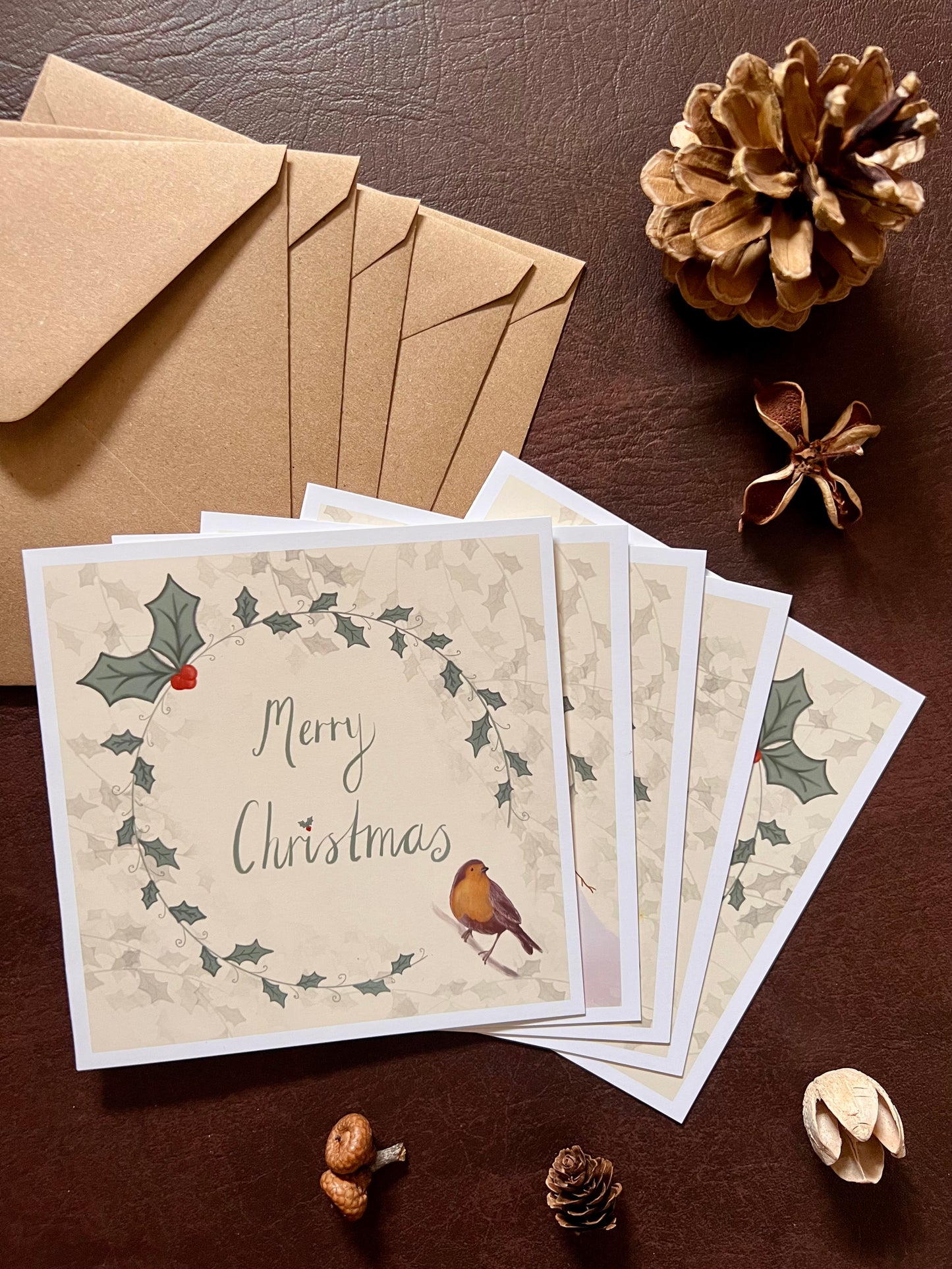 Flat lay image showing a stack of five artisan Christmas cards arranged in a fan shape on a tabletop with matching kraft envelopes. The robin design is on top, with other festive designs visible underneath including a snowman, reindeer, owl carrying a star, and a mouse decorating a Christmas tree, all featuring holly wreath borders and handwritten style 'Merry Christmas' text.