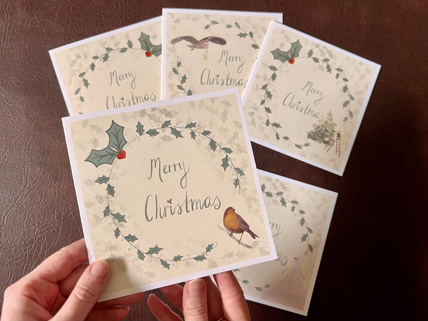 Hand holding the robin Christmas card design up towards the camera, featuring a holly wreath border and handwritten style 'Merry Christmas' text. The remaining cards from the five-card festive collection are laid flat on the tabletop below in the background.