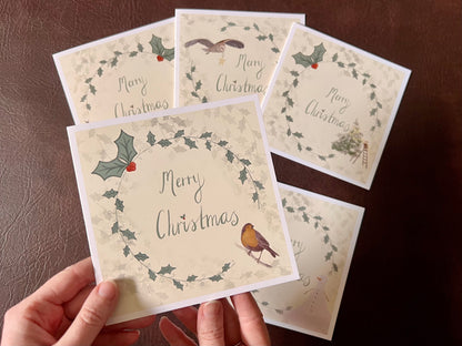 Hand holding the robin Christmas card design up towards the camera, featuring a holly wreath border and handwritten style 'Merry Christmas' text. The remaining cards from the five-card festive collection are laid flat on the tabletop below in the background.