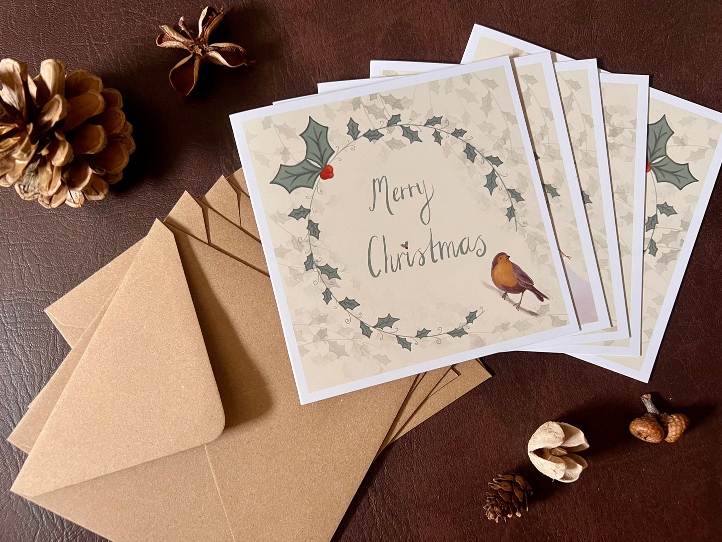 Flat lay image showing a pack of five artisan Christmas cards arranged in a fan shape on a leatherette tabletop with matching kraft envelopes. The collection includes traditional festive designs featuring a mouse decorating a Christmas tree, an owl carrying a star, a snowman, a reindeer and a robin, all with holly wreath borders and handwritten style 'Merry Christmas' text.