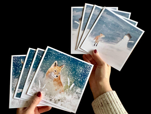 
Hands holding a pack of eight artisan Christmas cards arranged in a fan. The collection features winter wildlife and festive scenes including a fox, reindeer, deer, rabbit, snowman with fox cub, snowman with stag, snowman with rabbit, snowman with robin, and snowman with fox in snowy night settings.