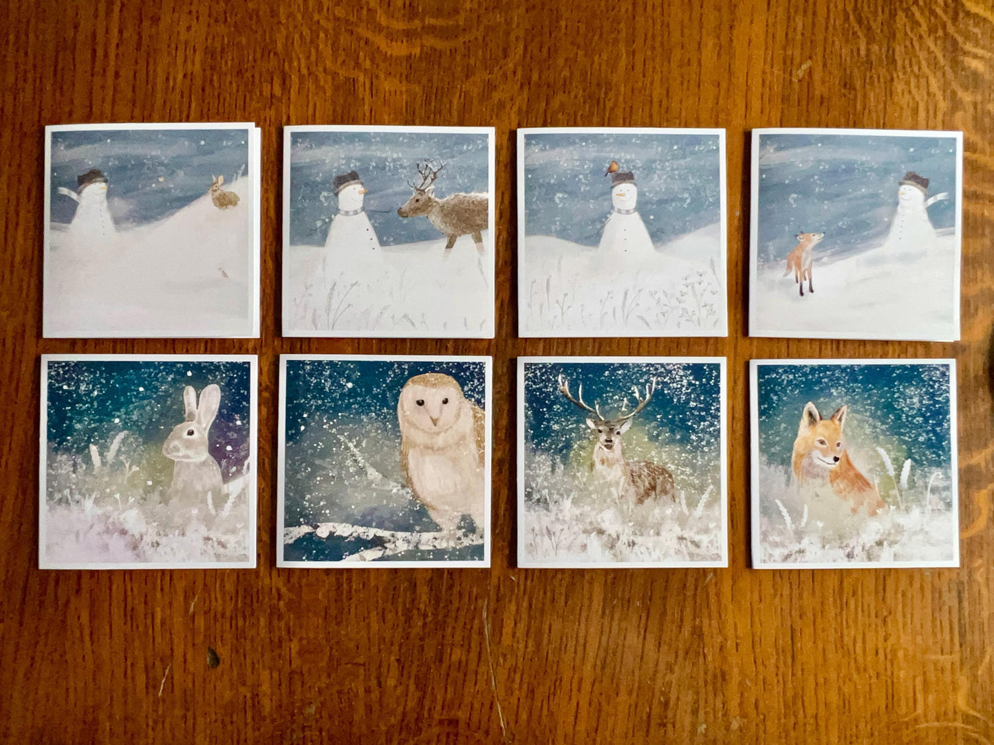 Flat lay image showing eight artisan Christmas cards arranged on a tabletop without their envelopes. The collection features winter wildlife and festive scenes including a fox, reindeer, deer, rabbit, snowman with fox cub, snowman with stag, snowman with rabbit, snowman with robin, and snowman with fox in snowy night settings.