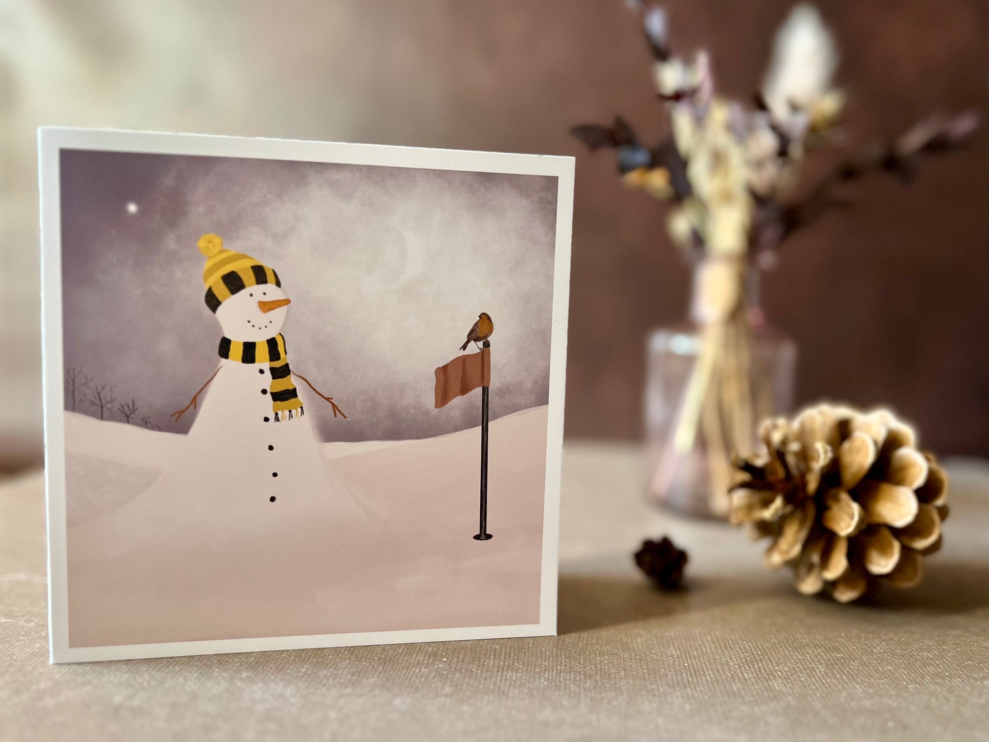 Artisan Christmas card featuring a snowman standing next to a golf flag on a snowy golf course with a robin perched on the flag, shown standing upright on a tabletop. The snowman is wearing a black and amber scarf and matching bobble hat in a sports inspired design.
