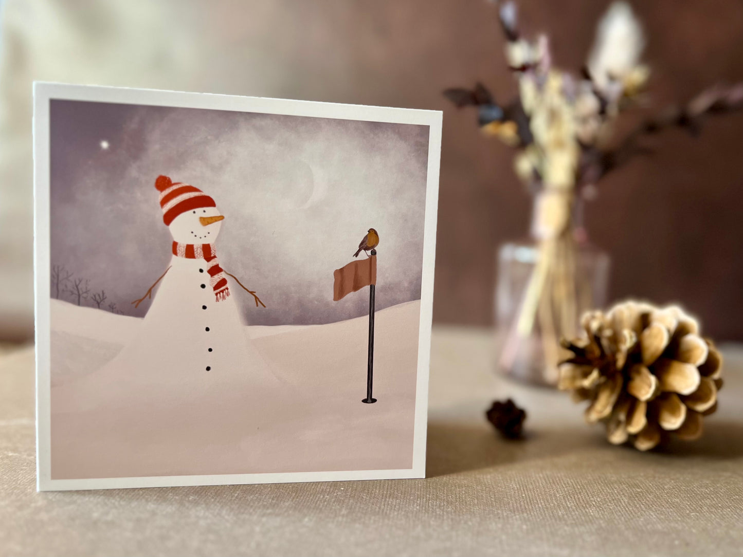 Artisan Christmas card featuring a snowman standing next to a golf flag on a snowy golf course with a robin perched on the flag, shown standing upright on a tabletop. The snowman is wearing a red and white scarf and matching bobble hat in a festive sports inspired design.
