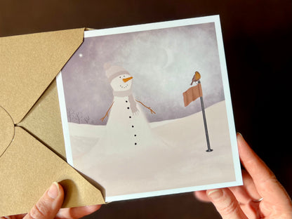 Hands holding an artisan Christmas card being removed from a kraft envelope, featuring a snowman standing next to a golf flag on a snowy golf course with a robin perched on the flag. The snowman is wearing a pale lilac and white scarf and matching bobble hat in a neutral winter sports inspired design.