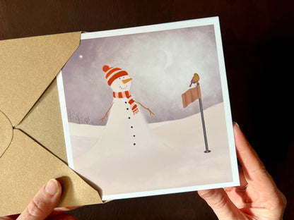 Hands holding an artisan Christmas card being removed from a kraft envelope, featuring a snowman standing next to a golf flag on a snowy golf course with a robin perched on the flag. The snowman is wearing a red and white scarf and matching bobble hat in a festive sports inspired design.