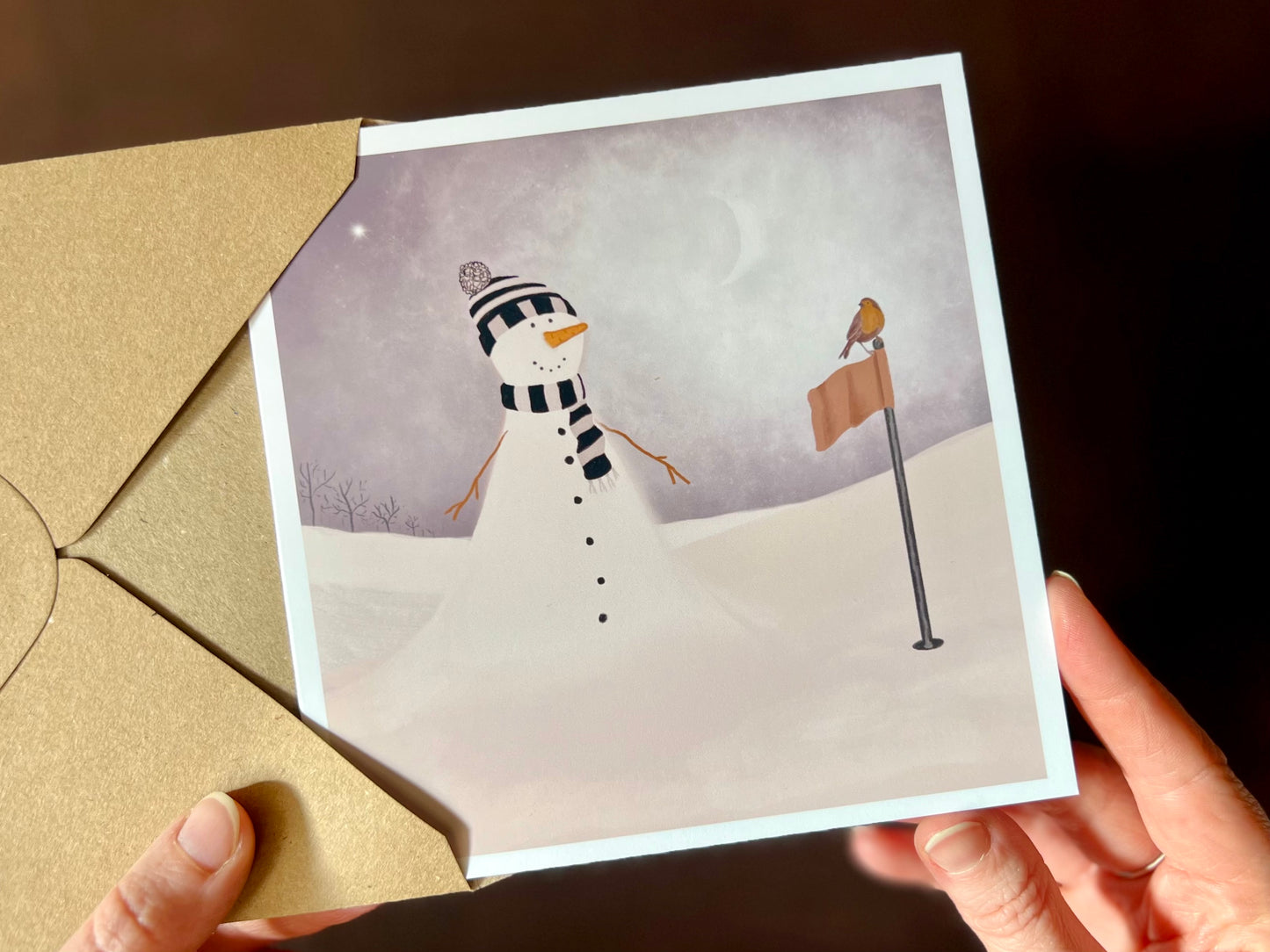 Hands holding an artisan Christmas card being removed from a kraft envelope, featuring a snowman standing next to a golf flag on a snowy golf course with a robin perched on the flag. The snowman is wearing a black and white striped scarf and matching bobble hat in a sports inspired design.