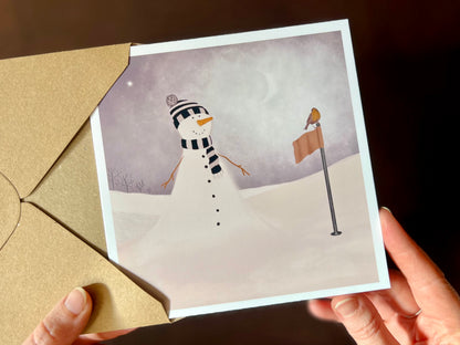 Hands holding an artisan Christmas card being removed from a kraft envelope, featuring a snowman standing next to a golf flag on a snowy golf course with a robin perched on the flag. The snowman is wearing a black and white striped scarf and matching bobble hat in a sports inspired design.